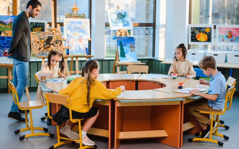 Dzieci przy wspólnym stole podczas zajęć, prowadzący obserwuje pracę